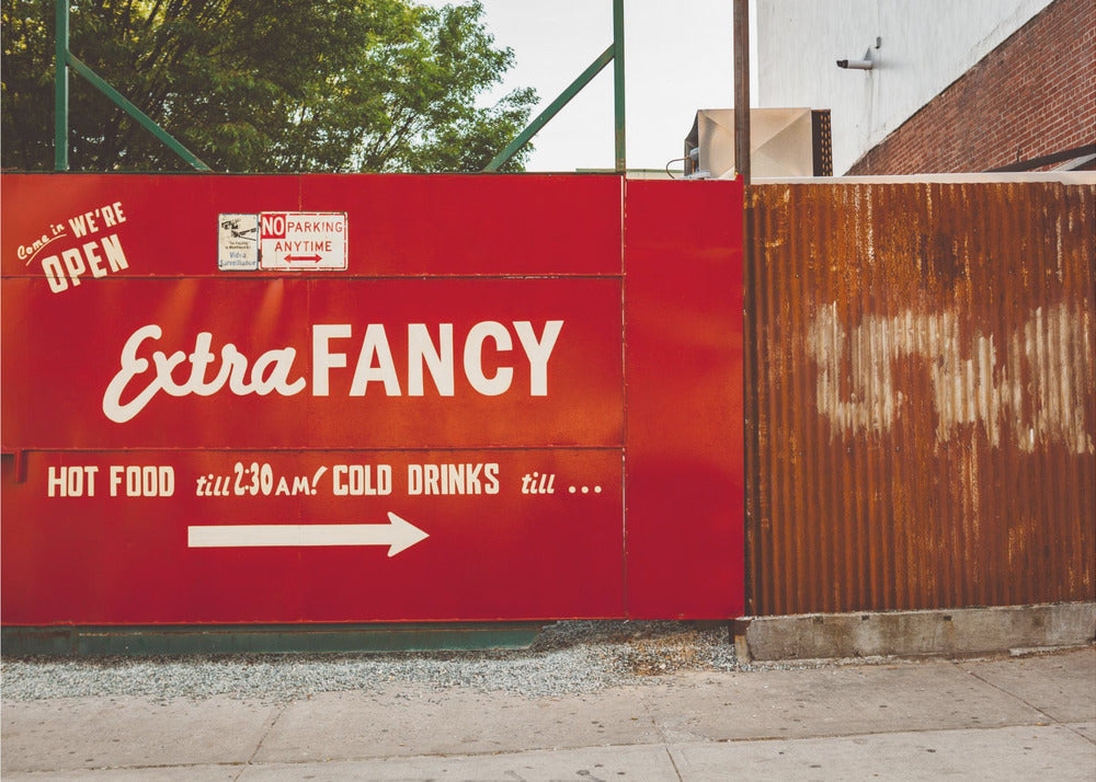 A framed photograph of a bright red outdoor sign for a restaurant named 'Extra Fancy'. The sign, painted with white retro-style text, advertises hot food and cold drinks and is situated next to a rusty, corrugated metal fence on a city sidewalk. Artwork