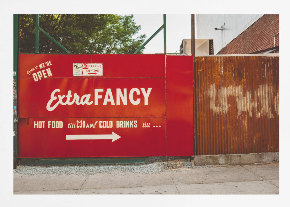 A framed photograph of a bright red outdoor sign for a restaurant named 'Extra Fancy'. The sign, painted with white retro-style text, advertises hot food and cold drinks and is situated next to a rusty, corrugated metal fence on a city sidewalk. Artwork