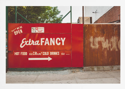 A framed photograph of a bright red outdoor sign for a restaurant named 'Extra Fancy'. The sign, painted with white retro-style text, advertises hot food and cold drinks and is situated next to a rusty, corrugated metal fence on a city sidewalk. Artwork