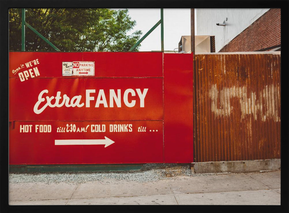 A framed photograph of a bright red outdoor sign for a restaurant named 'Extra Fancy'. The sign, painted with white retro-style text, advertises hot food and cold drinks and is situated next to a rusty, corrugated metal fence on a city sidewalk. Artwork
