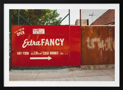 A framed photograph of a bright red outdoor sign for a restaurant named 'Extra Fancy'. The sign, painted with white retro-style text, advertises hot food and cold drinks and is situated next to a rusty, corrugated metal fence on a city sidewalk. Artwork