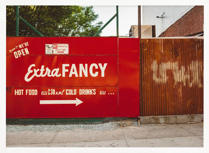 A framed photograph of a bright red outdoor sign for a restaurant named 'Extra Fancy'. The sign, painted with white retro-style text, advertises hot food and cold drinks and is situated next to a rusty, corrugated metal fence on a city sidewalk. Artwork