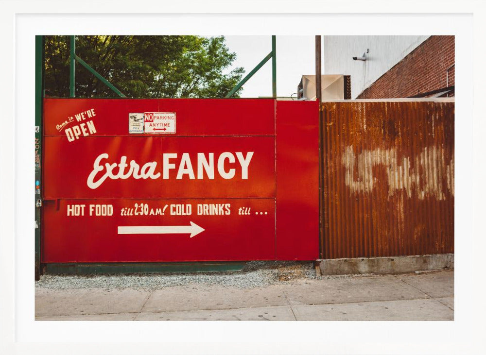 A framed photograph of a bright red outdoor sign for a restaurant named 'Extra Fancy'. The sign, painted with white retro-style text, advertises hot food and cold drinks and is situated next to a rusty, corrugated metal fence on a city sidewalk. Artwork