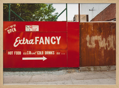 A framed photograph of a bright red outdoor sign for a restaurant named 'Extra Fancy'. The sign, painted with white retro-style text, advertises hot food and cold drinks and is situated next to a rusty, corrugated metal fence on a city sidewalk. Artwork