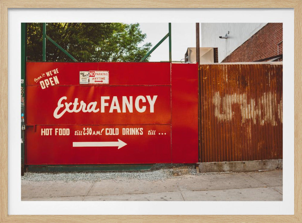 A framed photograph of a bright red outdoor sign for a restaurant named 'Extra Fancy'. The sign, painted with white retro-style text, advertises hot food and cold drinks and is situated next to a rusty, corrugated metal fence on a city sidewalk. Artwork