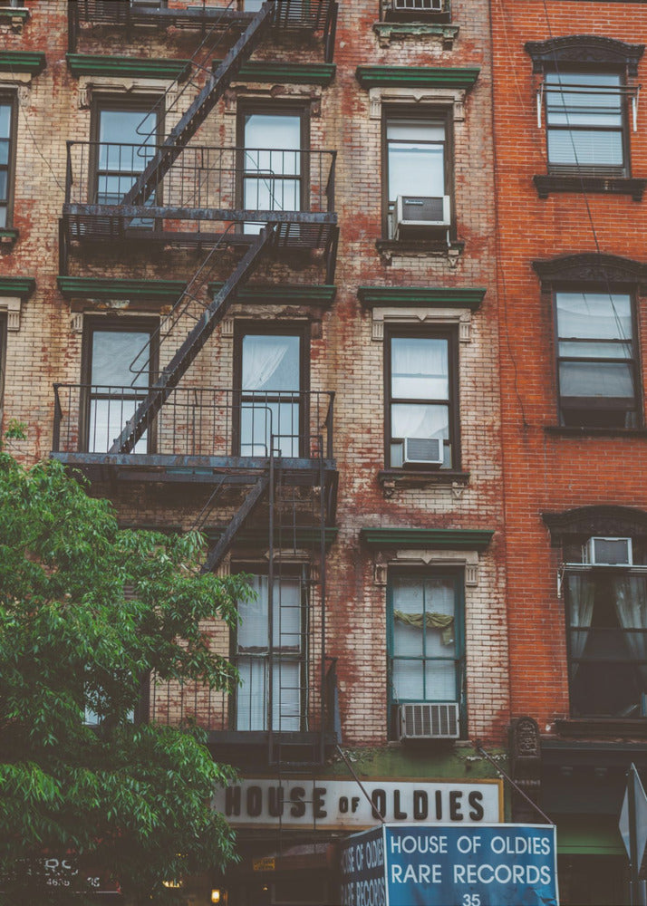 A classic multi-story brick apartment building with a prominent black metal fire escape zigzagging across its facade. A sign for a 'House of Oldies' rare records shop is visible on the ground floor. Poster
