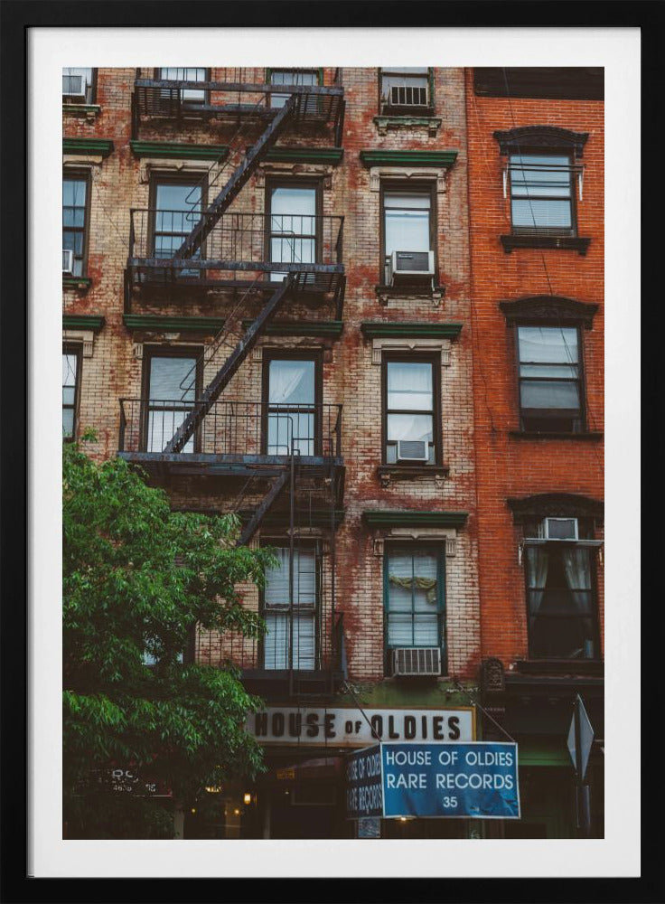 A classic multi-story brick apartment building with a prominent black metal fire escape zigzagging across its facade. A sign for a 'House of Oldies' rare records shop is visible on the ground floor. Poster