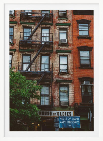 A classic multi-story brick apartment building with a prominent black metal fire escape zigzagging across its facade. A sign for a 'House of Oldies' rare records shop is visible on the ground floor. Poster