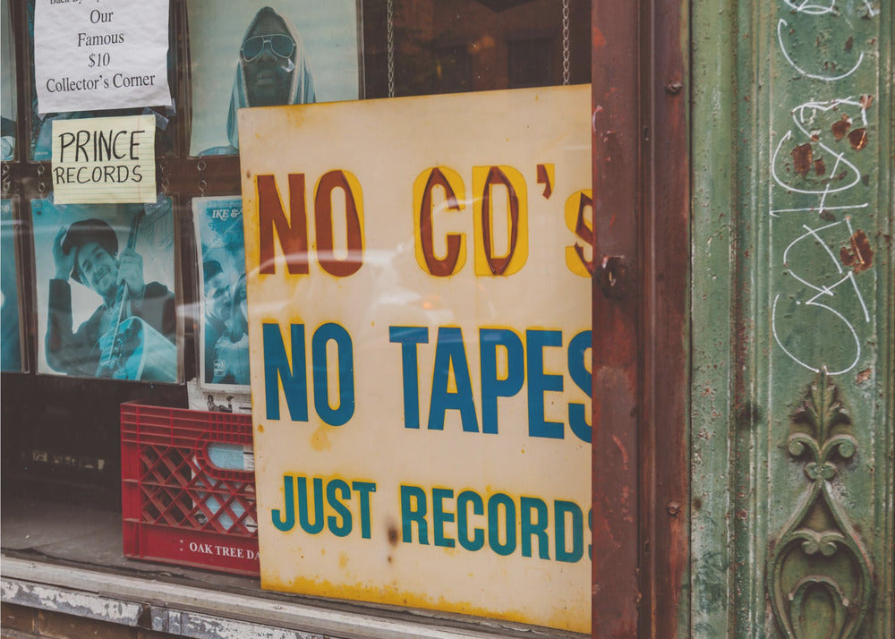 A weathered yellow sign in a record store window reads 'NO CD'S NO TAPES JUST RECORDS' in bold lettering. Behind the sign, vintage album covers and other posters are visible in the shop's display, next to a rustic green and brown door frame. Decor