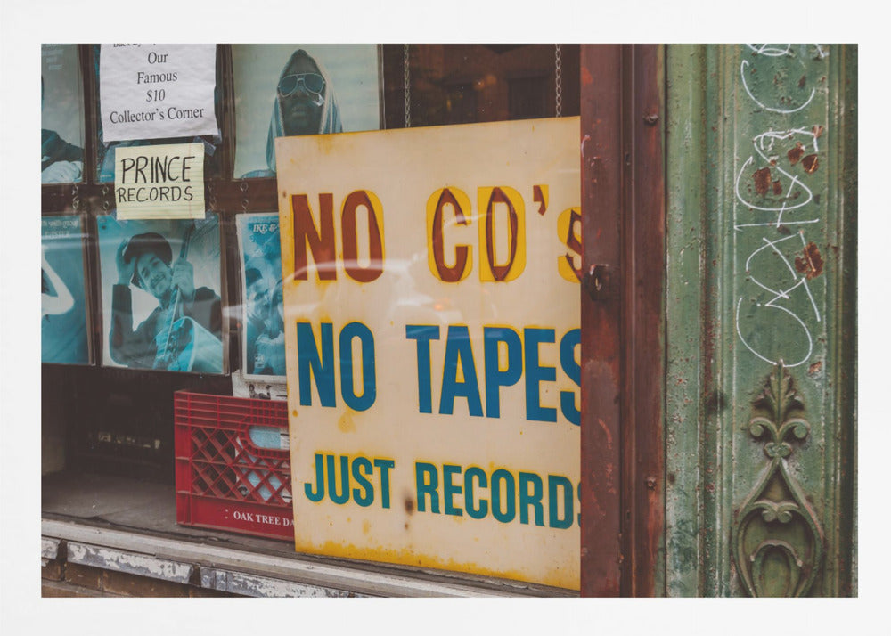 A weathered yellow sign in a record store window reads 'NO CD'S NO TAPES JUST RECORDS' in bold lettering. Behind the sign, vintage album covers and other posters are visible in the shop's display, next to a rustic green and brown door frame. Decor