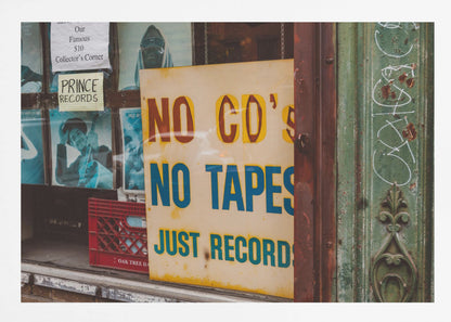 A weathered yellow sign in a record store window reads 'NO CD'S NO TAPES JUST RECORDS' in bold lettering. Behind the sign, vintage album covers and other posters are visible in the shop's display, next to a rustic green and brown door frame. Decor