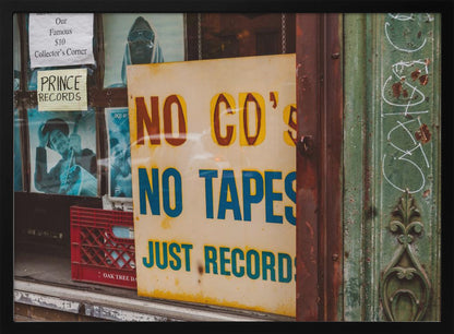 A weathered yellow sign in a record store window reads 'NO CD'S NO TAPES JUST RECORDS' in bold lettering. Behind the sign, vintage album covers and other posters are visible in the shop's display, next to a rustic green and brown door frame. Decor