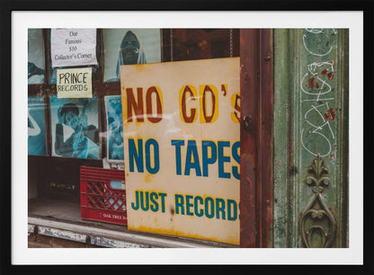 A weathered yellow sign in a record store window reads 'NO CD'S NO TAPES JUST RECORDS' in bold lettering. Behind the sign, vintage album covers and other posters are visible in the shop's display, next to a rustic green and brown door frame. Decor