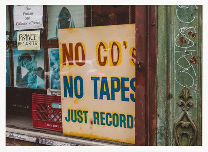 A weathered yellow sign in a record store window reads 'NO CD'S NO TAPES JUST RECORDS' in bold lettering. Behind the sign, vintage album covers and other posters are visible in the shop's display, next to a rustic green and brown door frame. Decor