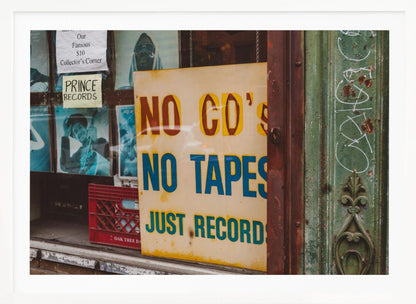 A weathered yellow sign in a record store window reads 'NO CD'S NO TAPES JUST RECORDS' in bold lettering. Behind the sign, vintage album covers and other posters are visible in the shop's display, next to a rustic green and brown door frame. Decor