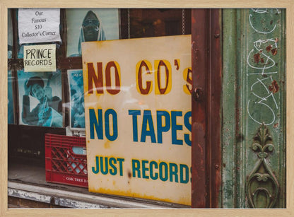 A weathered yellow sign in a record store window reads 'NO CD'S NO TAPES JUST RECORDS' in bold lettering. Behind the sign, vintage album covers and other posters are visible in the shop's display, next to a rustic green and brown door frame. Decor