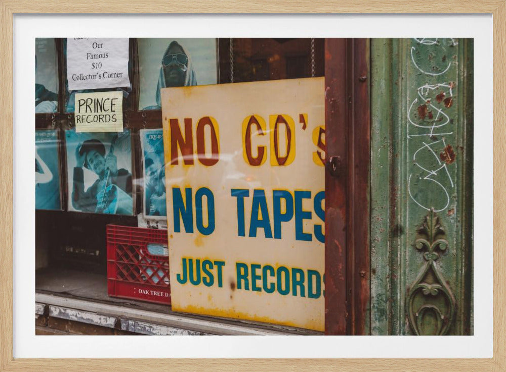 A weathered yellow sign in a record store window reads 'NO CD'S NO TAPES JUST RECORDS' in bold lettering. Behind the sign, vintage album covers and other posters are visible in the shop's display, next to a rustic green and brown door frame. Decor