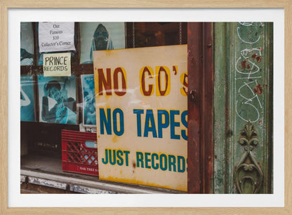A weathered yellow sign in a record store window reads 'NO CD'S NO TAPES JUST RECORDS' in bold lettering. Behind the sign, vintage album covers and other posters are visible in the shop's display, next to a rustic green and brown door frame. Decor