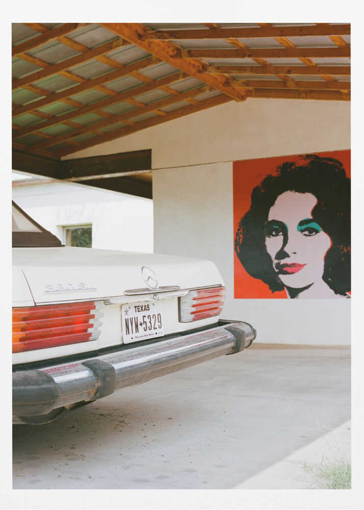 A film-style photograph showing the rear of a classic white Mercedes-Benz parked under a carport with exposed wooden beams. On the adjacent white wall hangs a vibrant red pop-art portrait of a woman with dark hair. Artwork