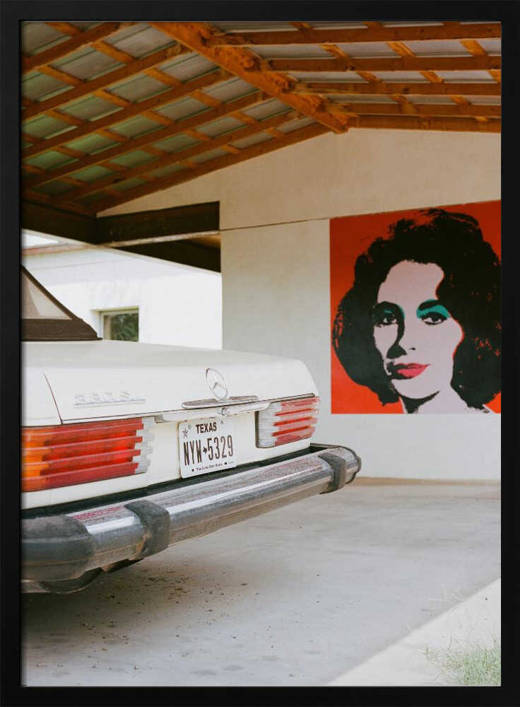 A film-style photograph showing the rear of a classic white Mercedes-Benz parked under a carport with exposed wooden beams. On the adjacent white wall hangs a vibrant red pop-art portrait of a woman with dark hair. Artwork