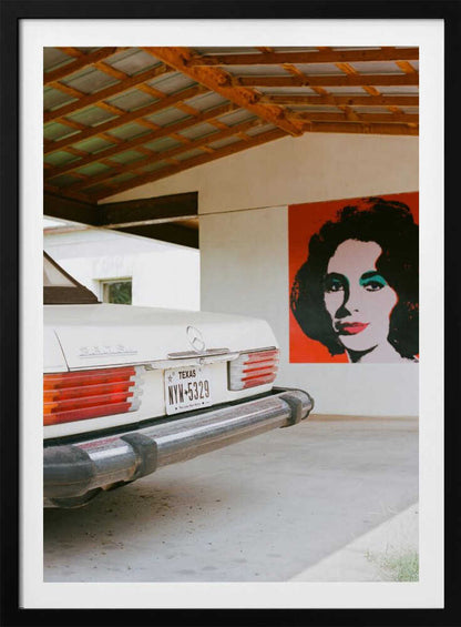 A film-style photograph showing the rear of a classic white Mercedes-Benz parked under a carport with exposed wooden beams. On the adjacent white wall hangs a vibrant red pop-art portrait of a woman with dark hair. Artwork