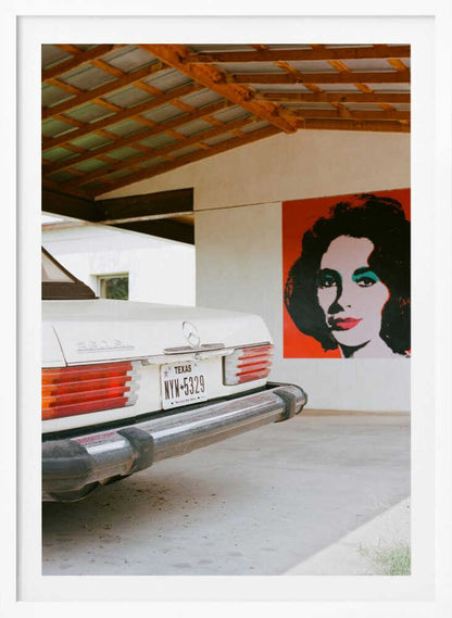 A film-style photograph showing the rear of a classic white Mercedes-Benz parked under a carport with exposed wooden beams. On the adjacent white wall hangs a vibrant red pop-art portrait of a woman with dark hair. Artwork
