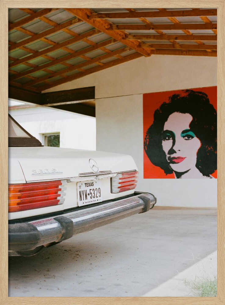 A film-style photograph showing the rear of a classic white Mercedes-Benz parked under a carport with exposed wooden beams. On the adjacent white wall hangs a vibrant red pop-art portrait of a woman with dark hair. Artwork