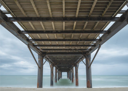 A symmetrical, low-angle photograph taken from underneath a long pier, looking out to sea. The rusty metal pilings and wooden planks of the pier create a receding perspective over the smooth, long-exposure water, all under a cloudy sky and presented within a silver frame. Poster