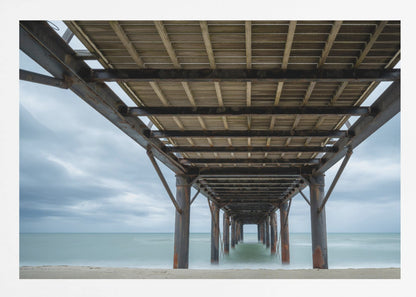 A symmetrical, low-angle photograph taken from underneath a long pier, looking out to sea. The rusty metal pilings and wooden planks of the pier create a receding perspective over the smooth, long-exposure water, all under a cloudy sky and presented within a silver frame. Poster