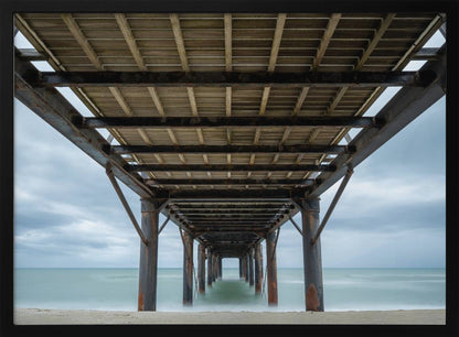 A symmetrical, low-angle photograph taken from underneath a long pier, looking out to sea. The rusty metal pilings and wooden planks of the pier create a receding perspective over the smooth, long-exposure water, all under a cloudy sky and presented within a silver frame. Poster