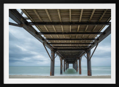 A symmetrical, low-angle photograph taken from underneath a long pier, looking out to sea. The rusty metal pilings and wooden planks of the pier create a receding perspective over the smooth, long-exposure water, all under a cloudy sky and presented within a silver frame. Poster