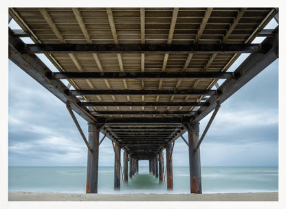 A symmetrical, low-angle photograph taken from underneath a long pier, looking out to sea. The rusty metal pilings and wooden planks of the pier create a receding perspective over the smooth, long-exposure water, all under a cloudy sky and presented within a silver frame. Poster