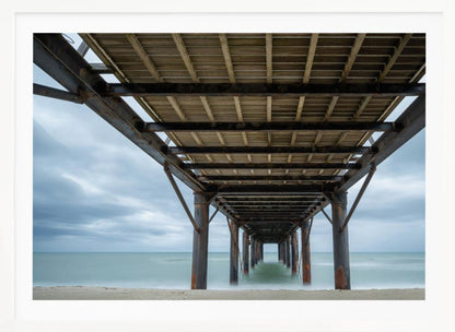 A symmetrical, low-angle photograph taken from underneath a long pier, looking out to sea. The rusty metal pilings and wooden planks of the pier create a receding perspective over the smooth, long-exposure water, all under a cloudy sky and presented within a silver frame. Poster