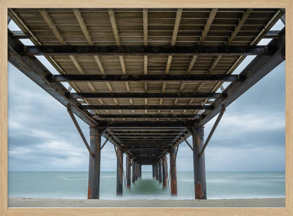 A symmetrical, low-angle photograph taken from underneath a long pier, looking out to sea. The rusty metal pilings and wooden planks of the pier create a receding perspective over the smooth, long-exposure water, all under a cloudy sky and presented within a silver frame. Poster