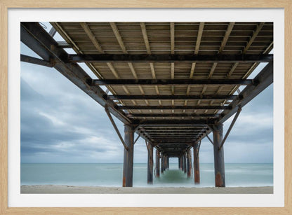 A symmetrical, low-angle photograph taken from underneath a long pier, looking out to sea. The rusty metal pilings and wooden planks of the pier create a receding perspective over the smooth, long-exposure water, all under a cloudy sky and presented within a silver frame. Poster
