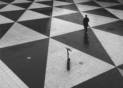 A high-angle, black and white photograph of a man in a coat and fedora walking away across a vast plaza with a geometric pattern of black and light gray triangles. An electric scooter stands alone in the foreground, creating a sense of solitude and contrast. The image is presented within a silver frame. Decor