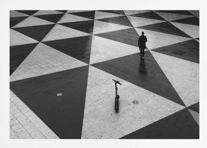 A high-angle, black and white photograph of a man in a coat and fedora walking away across a vast plaza with a geometric pattern of black and light gray triangles. An electric scooter stands alone in the foreground, creating a sense of solitude and contrast. The image is presented within a silver frame. Decor