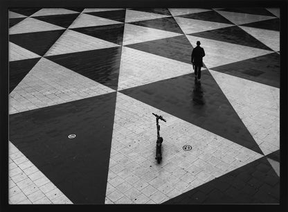 A high-angle, black and white photograph of a man in a coat and fedora walking away across a vast plaza with a geometric pattern of black and light gray triangles. An electric scooter stands alone in the foreground, creating a sense of solitude and contrast. The image is presented within a silver frame. Decor