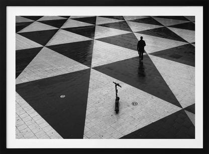 A high-angle, black and white photograph of a man in a coat and fedora walking away across a vast plaza with a geometric pattern of black and light gray triangles. An electric scooter stands alone in the foreground, creating a sense of solitude and contrast. The image is presented within a silver frame. Decor