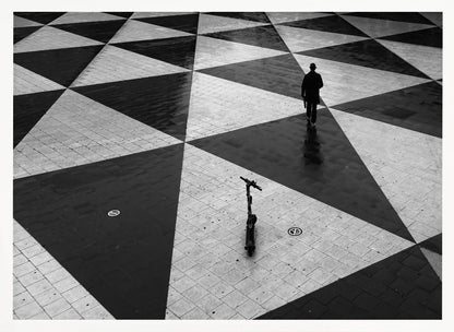 A high-angle, black and white photograph of a man in a coat and fedora walking away across a vast plaza with a geometric pattern of black and light gray triangles. An electric scooter stands alone in the foreground, creating a sense of solitude and contrast. The image is presented within a silver frame. Decor