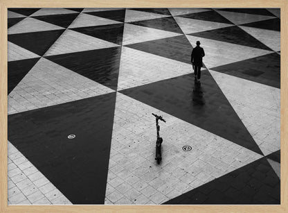 A high-angle, black and white photograph of a man in a coat and fedora walking away across a vast plaza with a geometric pattern of black and light gray triangles. An electric scooter stands alone in the foreground, creating a sense of solitude and contrast. The image is presented within a silver frame. Decor