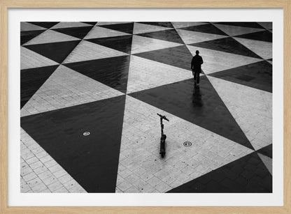 A high-angle, black and white photograph of a man in a coat and fedora walking away across a vast plaza with a geometric pattern of black and light gray triangles. An electric scooter stands alone in the foreground, creating a sense of solitude and contrast. The image is presented within a silver frame. Decor