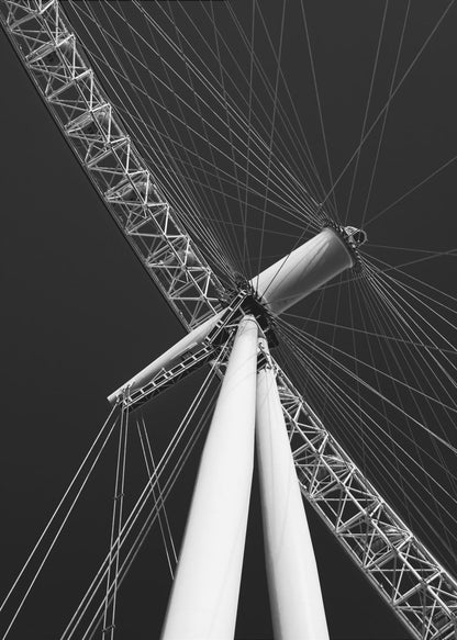 A dramatic low-angle, black and white photograph of a large Ferris wheel, focusing on the central hub and supporting structure. The image captures the white A-frame legs and the intricate lattice of the wheel's rim, with numerous tension cables radiating outwards against a solid black sky, creating a strong sense of perspective and geometric pattern. Artwork