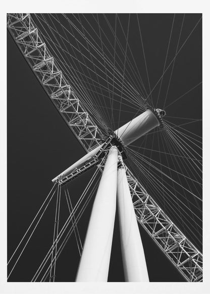 A dramatic low-angle, black and white photograph of a large Ferris wheel, focusing on the central hub and supporting structure. The image captures the white A-frame legs and the intricate lattice of the wheel's rim, with numerous tension cables radiating outwards against a solid black sky, creating a strong sense of perspective and geometric pattern. Artwork