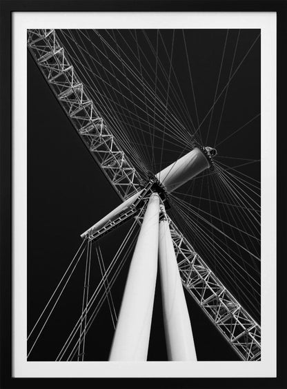 A dramatic low-angle, black and white photograph of a large Ferris wheel, focusing on the central hub and supporting structure. The image captures the white A-frame legs and the intricate lattice of the wheel's rim, with numerous tension cables radiating outwards against a solid black sky, creating a strong sense of perspective and geometric pattern. Artwork