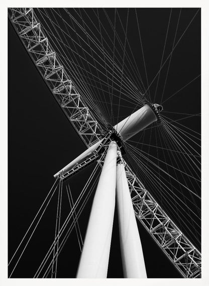 A dramatic low-angle, black and white photograph of a large Ferris wheel, focusing on the central hub and supporting structure. The image captures the white A-frame legs and the intricate lattice of the wheel's rim, with numerous tension cables radiating outwards against a solid black sky, creating a strong sense of perspective and geometric pattern. Artwork