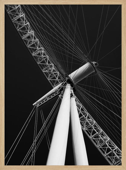 A dramatic low-angle, black and white photograph of a large Ferris wheel, focusing on the central hub and supporting structure. The image captures the white A-frame legs and the intricate lattice of the wheel's rim, with numerous tension cables radiating outwards against a solid black sky, creating a strong sense of perspective and geometric pattern. Artwork
