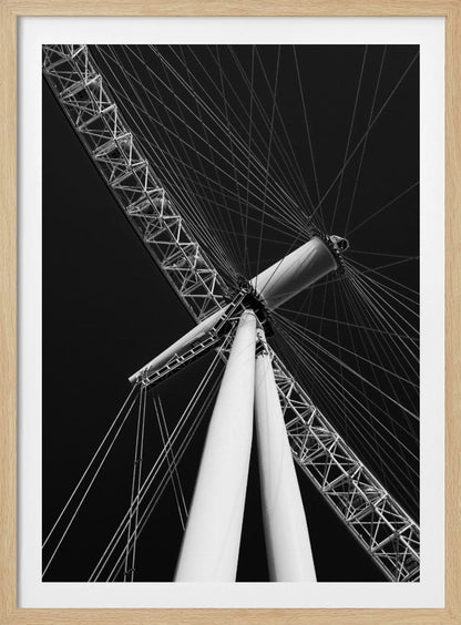 A dramatic low-angle, black and white photograph of a large Ferris wheel, focusing on the central hub and supporting structure. The image captures the white A-frame legs and the intricate lattice of the wheel's rim, with numerous tension cables radiating outwards against a solid black sky, creating a strong sense of perspective and geometric pattern. Artwork