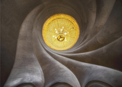 A worm's-eye view of an ornate, golden chandelier set against a swirling, textured dark grey ceiling. The spiraling architectural feature draws the eye to the brightly lit fixture, creating an abstract and dramatic composition. Poster