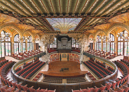 A high-angle, wide shot of the ornate and lavish interior of the Palau de la Música Catalana in Barcelona. The view captures the entire concert hall, with its curved balconies filled with red seats, a central wooden stage with a grand piano, and a spectacular, large stained-glass skylight in the ceiling. Poster